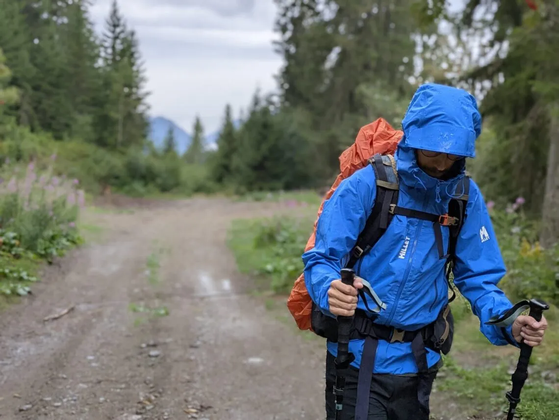 Randonneur en veste Millet Kamet Light bleue marchant sous la pluie sur une piste forestière