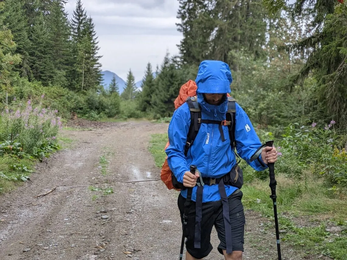 Randonneur en veste Millet Kamet Light remontant une piste détrempée sous la pluie