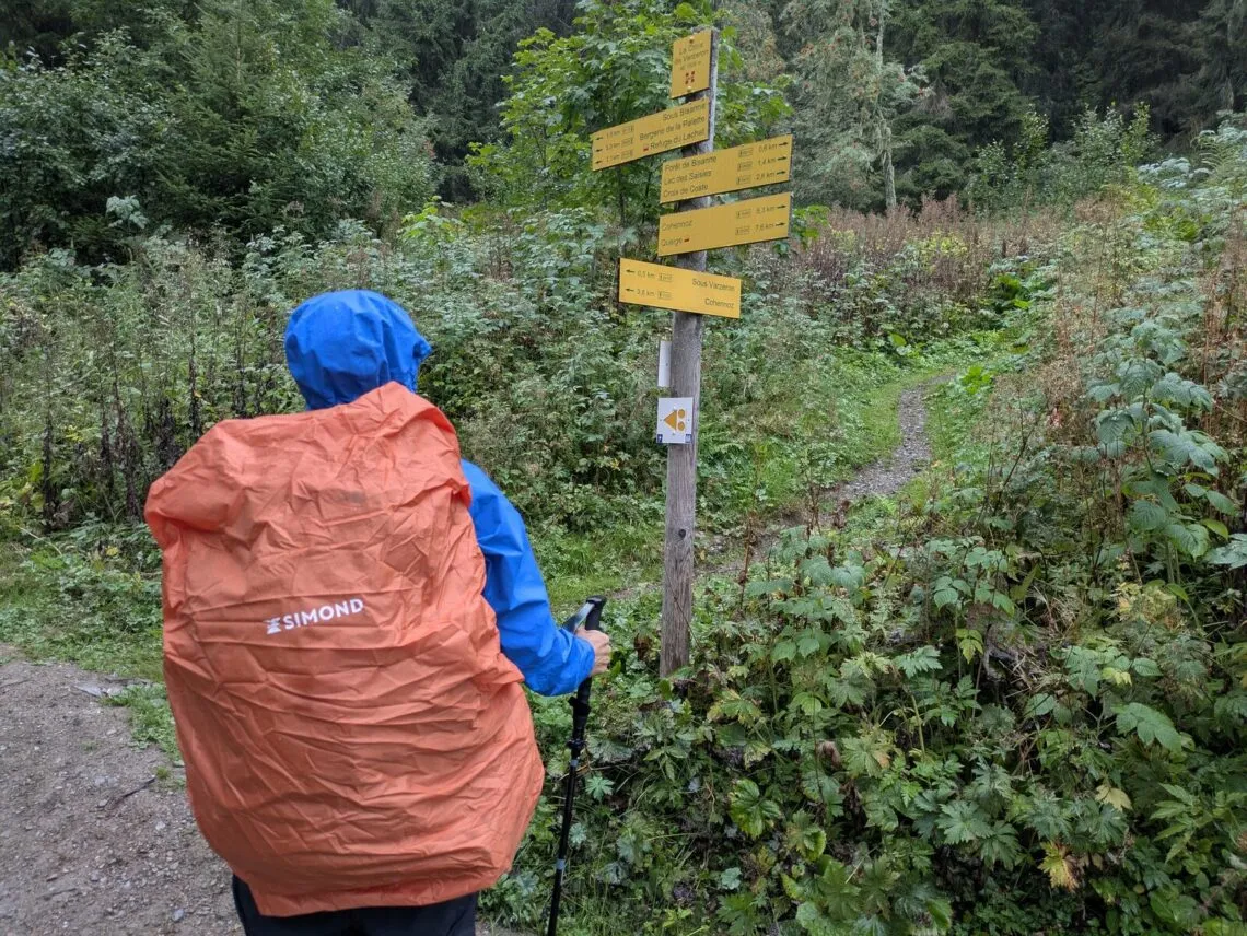 Randonneur de dos en veste Millet Kamet Light, sac couvert de sa housse de pluie, à une bifurcation du Tour du Beaufortain
